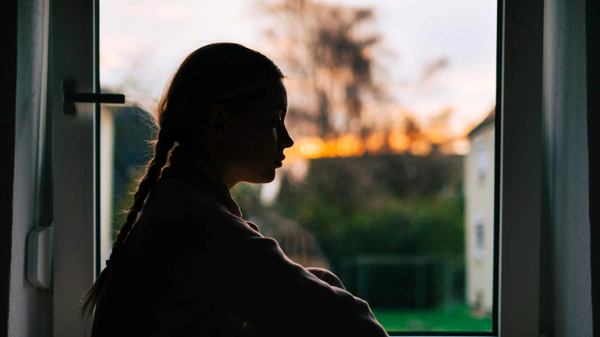 Fotografie zeigt Silhouette eines Mädchens mit geflochtenem Haar, das nachdenklich aus einem Fenster blickt. Hintergrund zeigt unscharfen Garten und Sonnenuntergang mit warmen Farben.