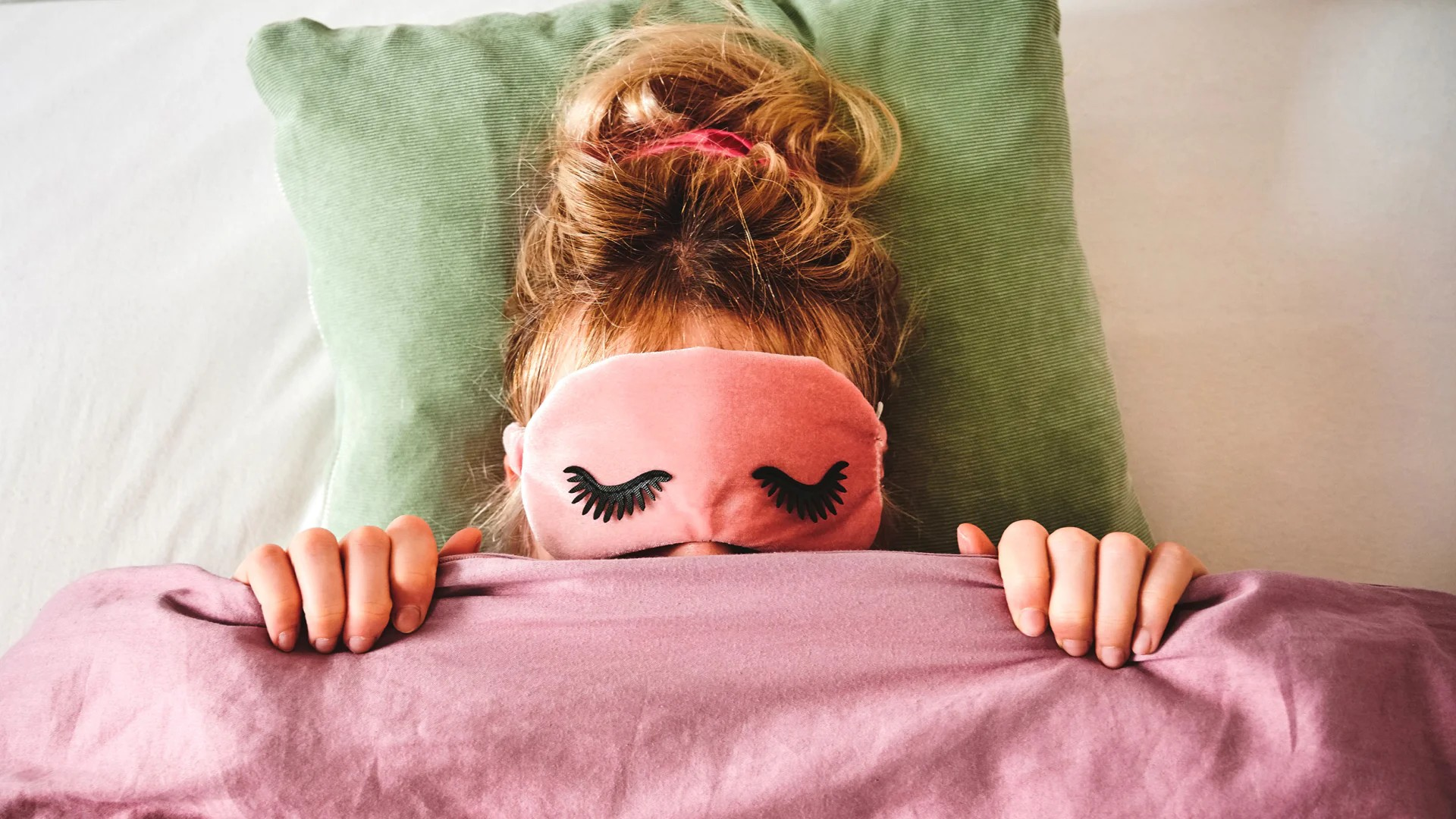 Fotografie einer Person mit rosa Schlafmaske und hochgestecktem Haar, die unter einer rosa Decke liegt und sich an deren Rand festhält. Hintergrund zeigt grünes Kissen und weiße Bettwäsche, vermittelt entspannte Schlafsituation.