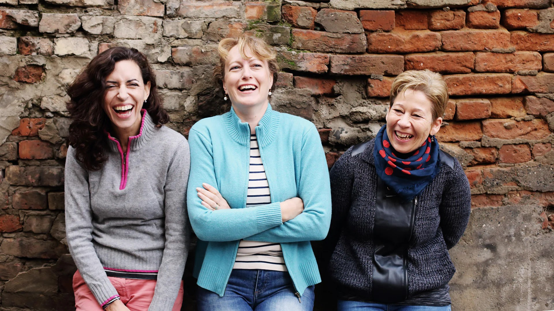 Fotografie von drei Frauen vor einer alten Backsteinmauer, die herzlich lachen und entspannt wirken. Die Frauen tragen lässige Kleidung in Grau, Hellblau und Dunkelblau mit einem Schal, was eine freundschaftliche und fröhliche Stimmung vermittelt.