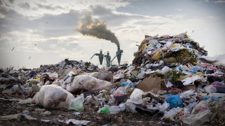 Fotografie einer großen Müllhalde mit Plastik- und Papierabfällen im Vordergrund und rauchenden Industrieanlagen im Hintergrund. Szene zeigt Umweltverschmutzung durch Abfallberge und Industrieemissionen unter bewölktem Himmel.