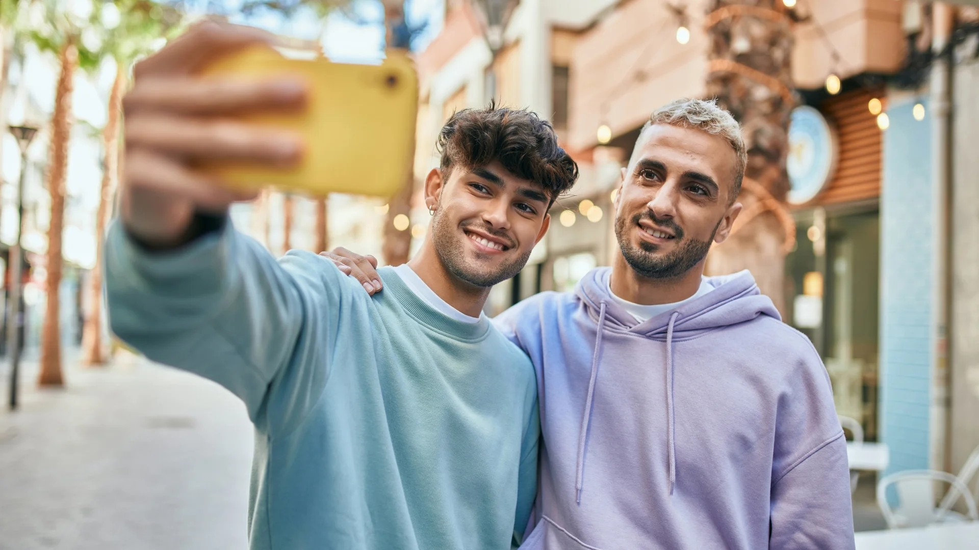 Zwei junge Männer auf einem Gehweg machen ein Selfie. Im Hintergrund Läden und LIcherketten