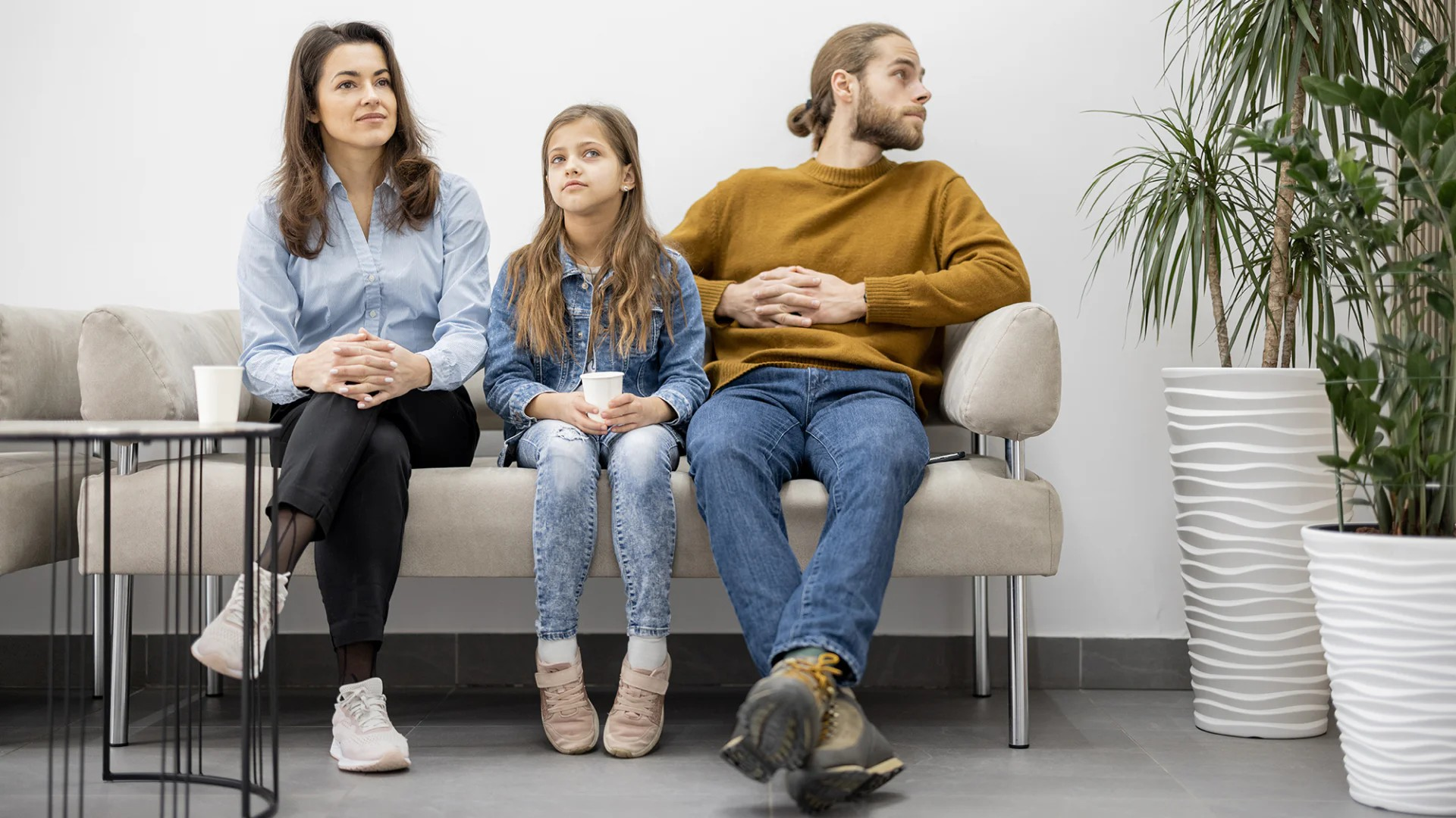 Fotografie zeigt drei Personen – Frau, Mädchen und Mann – auf Sofa sitzend, alle schauen in unterschiedliche Richtungen. Szene vermittelt unterschiedliche Stimmungen oder Gedanken, helle, moderne Umgebung mit Pflanzen und minimalistischer Einrichtung.