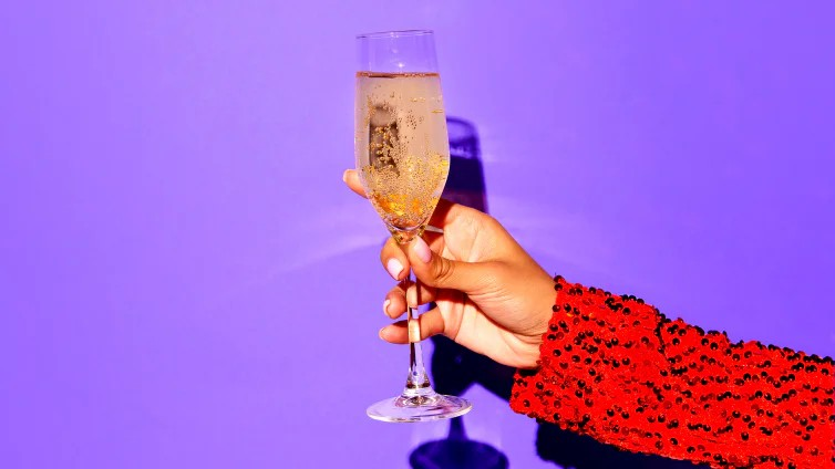 Fotografie einer Hand mit rotem, glitzerndem Ärmel, die ein gefülltes Champagnerglas vor lila Hintergrund hält. Fokus liegt auf Glas und Hand, Schattenwurf im Hintergrund betont Konturen und festliche Stimmung.